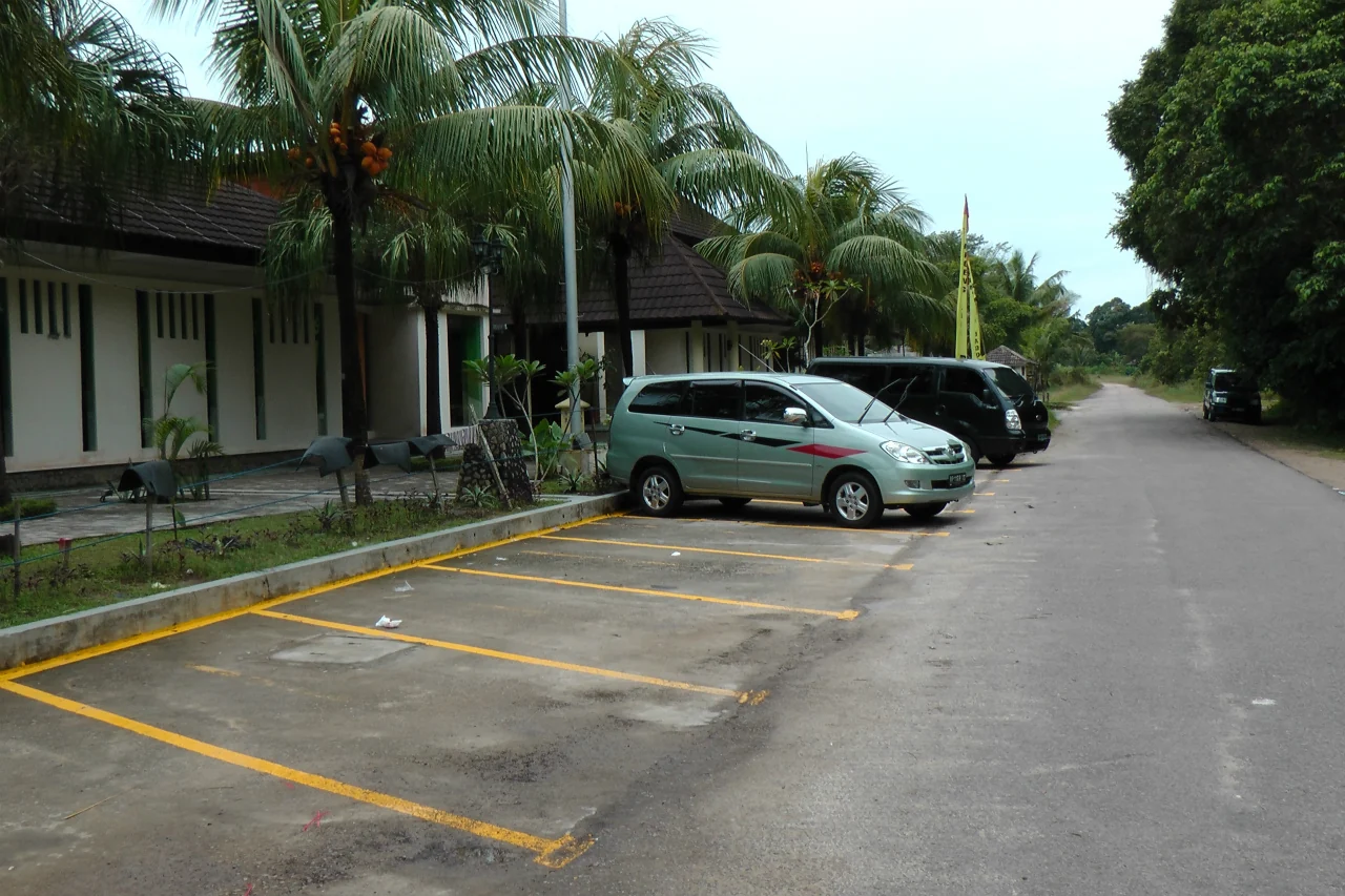 Jalur berpermukaan aspal, area parkir, bangunan rendah, dan pohon kelapa di Sebong Lagoi